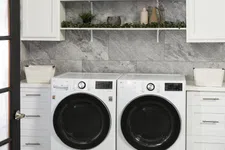 Thumbnail for Modern laundry room with front-load washer and dryer, white cabinetry, grey stone wall, and decorative shelves.