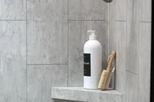 Thumbnail for White pump bottle and wooden brush on corner shelf in modern gray tiled shower area under black showerhead.
