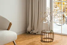 Thumbnail for Cozy corner with beige armchair, long curtains, round table with vase, and sunlight from window showing autumn trees.