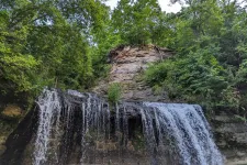 Thumbnail for Waterfall cascading over rocky cliffs surrounded by lush green forest under a clear sky.