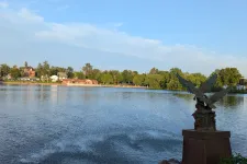Thumbnail for Eagle statue overlooking a calm lake with trees and houses under a clear blue sky at sunset.