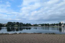 Thumbnail for Calm lake with reflections of trees and cloudy sky viewed from a brick-paved shoreline in a park.