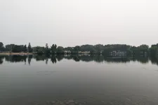 Thumbnail for Calm lake with clear water reflecting trees and overcast sky, shoreline visible with scattered rocks beneath water.