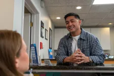 Thumbnail for Smiling man in plaid shirt talks to a woman across a reception desk in a professional office setting.