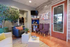 Thumbnail for Cozy waiting room with armchairs, two purple chairs, potted plants, bulletin board, TV, and wood-paneled wall.
