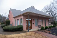 Thumbnail for Single-story red brick building with peaked roof and large wooden door surrounded by bushes and a paved walkway.