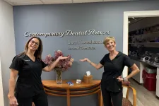 Thumbnail for Two smiling female dentists in black scrubs pose in front of Contemporary Dental Arts P.C. sign with flowers and brochures.