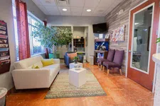 Thumbnail for Bright modern waiting room with beige sofa, purple chairs, large plant, TV, and decorative fish tank on wooden floor.