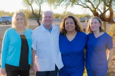 Thumbnail for Smiling healthcare team of four standing outdoors in scrubs and white coat with trees and car in background.
