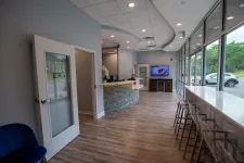 Thumbnail for Modern dental office reception area with curved stone desk, wood floor, and large windows with bar stools.