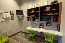 Thumbnail for Children's study area with green chairs, wall-mounted TV, shelves with toys, books, and kids' artwork on the wall.