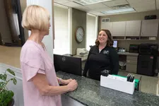Thumbnail for Receptionist smiling at a female visitor across a modern office front desk with computer and clock.