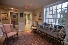 Thumbnail for Cozy waiting room with vintage armchairs, a wooden bench with patterned cushions, and a large window letting in natural light.