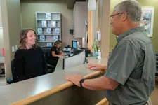 Thumbnail for Smiling receptionist assisting an older male client at a medical office front desk with a colleague working in the background