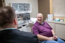 Thumbnail for Smiling man in purple shirt sitting in a medical office talking with a healthcare professional.