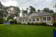 Thumbnail for Modern single-story building with stone and siding exterior surrounded by green lawn and trees under cloudy sky.
