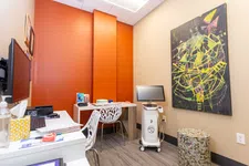 Thumbnail for Modern dental office with orange accent wall, white desks, abstract painting, and dental equipment on wheels.