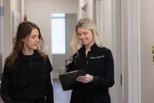 Thumbnail for Two women in black uniforms discussing notes on a clipboard in a bright medical office hallway.