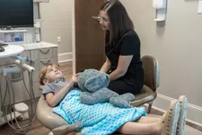 Thumbnail for Child lying on dental chair covered with blue blanket holding teddy bear while dentist talks to him