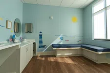 Thumbnail for Bright children's clinic room with blue nautical wall art, bench seating, white cabinets, and large window letting in sunlight
