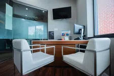 Thumbnail for Modern office with two white chairs facing a wooden desk, computer monitor, TV, and large window with blinds.