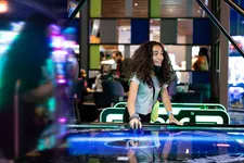 Thumbnail for Happy girl playing air hockey in a vibrant arcade, surrounded by colorful lights and excited players.