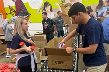 Thumbnail for Volunteers packing canned goods and jars into CSFP boxes on a conveyor in a food bank with mural background.