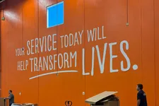 Thumbnail for Workers handle pallets and cardboard in a warehouse with an orange wall and overlaid text saying your service today will help transform lives.