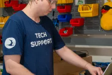Thumbnail for Tech support worker in blue shirt using laptop on a stack of boxes in a cluttered office workshop.