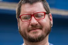 Thumbnail for Bearded man with red glasses and septum piercing wearing a blue Brutech t-shirt with arms crossed.