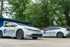Thumbnail for Two white Braintek business cars parked outside by blue building and green trees in daylight