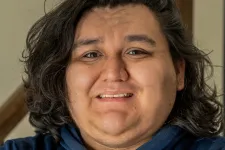 Thumbnail for Smiling young man with long dark hair wearing a blue hoodie stands with arms crossed indoors near stairs.
