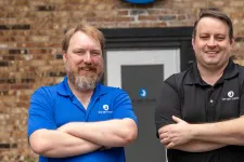 Thumbnail for Two smiling men in branded polo shirts standing with arms crossed outside a brick building with company logo.