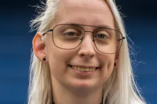 Thumbnail for Young woman with long blonde hair in glasses wearing a blue Braintek polo shirt smiling outdoors.