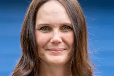 Thumbnail for Smiling woman with long brown hair wearing a black Braintek polo shirt with a blue background.