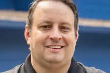 Thumbnail for Smiling man in black Braintek polo shirt with arms crossed against blue background.