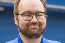 Thumbnail for Smiling man with glasses and beard wearing a blue Braintek polo shirt outdoors with a blurred blue background.