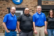 Thumbnail for Four employees in blue and black company polo shirts standing outdoors in front of a brick building with a blue logo on the wall.
