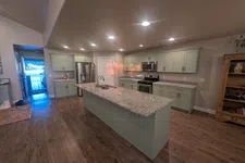 Thumbnail for Modern kitchen with large granite island, light green cabinets, stainless steel appliances, and wood flooring.