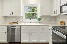 Thumbnail for Modern white kitchen with stainless steel appliances, black hardware, and a window overlooking green trees.