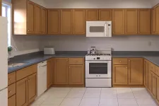 Thumbnail for Modern kitchen with wooden cabinets, white appliances, gray countertops, and tiled floor under bright lighting.