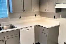 Thumbnail for Modern kitchen corner with gray cabinets, white countertop, dishwasher, stainless steel sink, and wood laminate floor.