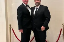 Thumbnail for Two men in black tuxedos posing in front of a Beverly Hilton wall with red velvet ropes around them.