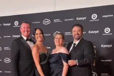 Thumbnail for Four people dressed in formal black attire posing on a red carpet in front of a branded backdrop at an event.