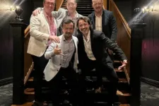 Thumbnail for Five men in formal attire posing on wooden staircase inside a dimly lit elegant building with carpeted floor.