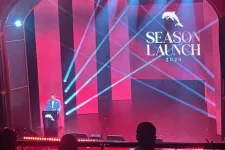 Thumbnail for Speaker at podium on stage with red panels and blue spotlight beams during 2026 Season Launch event