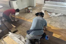 Thumbnail for Two workers installing dark wood flooring on a kitchen floor with tools and supplies around them.