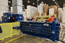 Thumbnail for Industrial paper baler and large rolls of paper stacked in a warehouse with equipment and safety barriers.