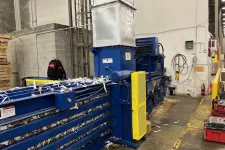 Thumbnail for Large blue industrial baler machine compressing shredded paper inside a warehouse with concrete walls and yellow safety barriers.