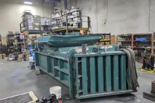 Thumbnail for Large industrial green metal baler machine inside a cluttered warehouse with shelves and boxes.